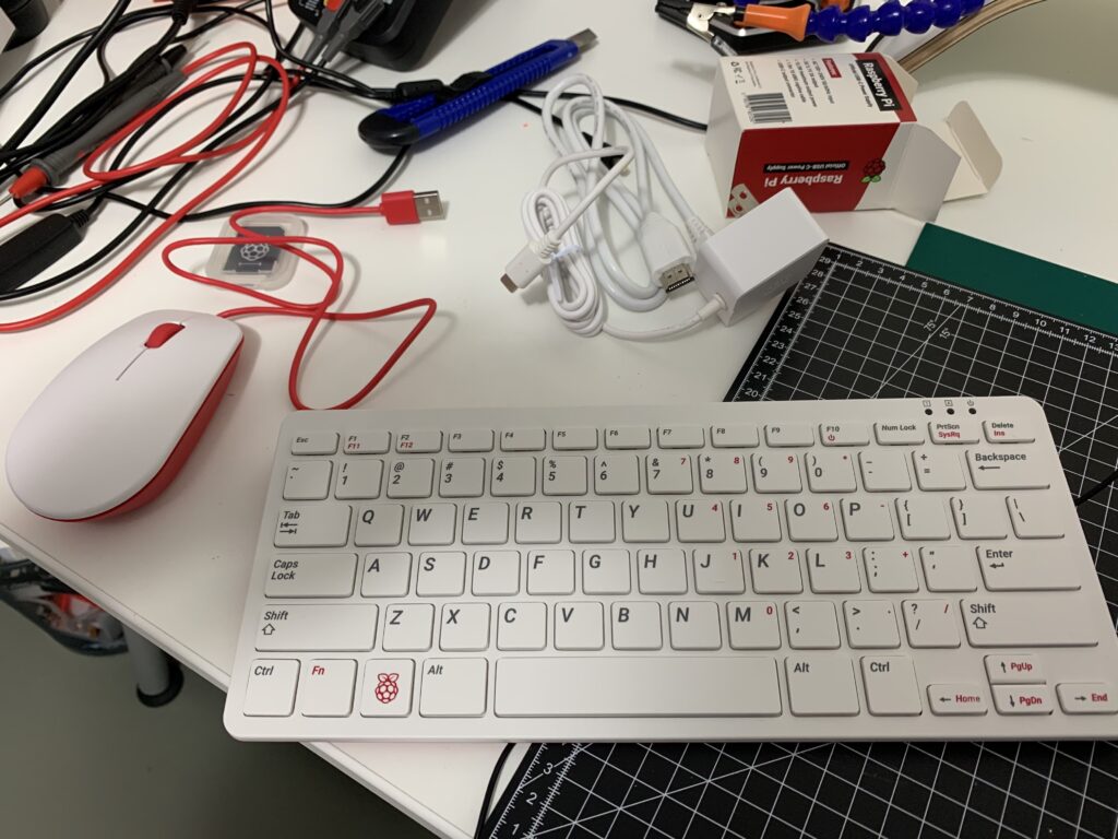 Foto com um computador RaspberryPi 400 na mesa. O computador vem montado em um case com o teclado. O teclado é branco com detalhes em preto e rosa. Do lado do computador tem um mouse branco e rosa e um monte de cabos que acompanham o kit.