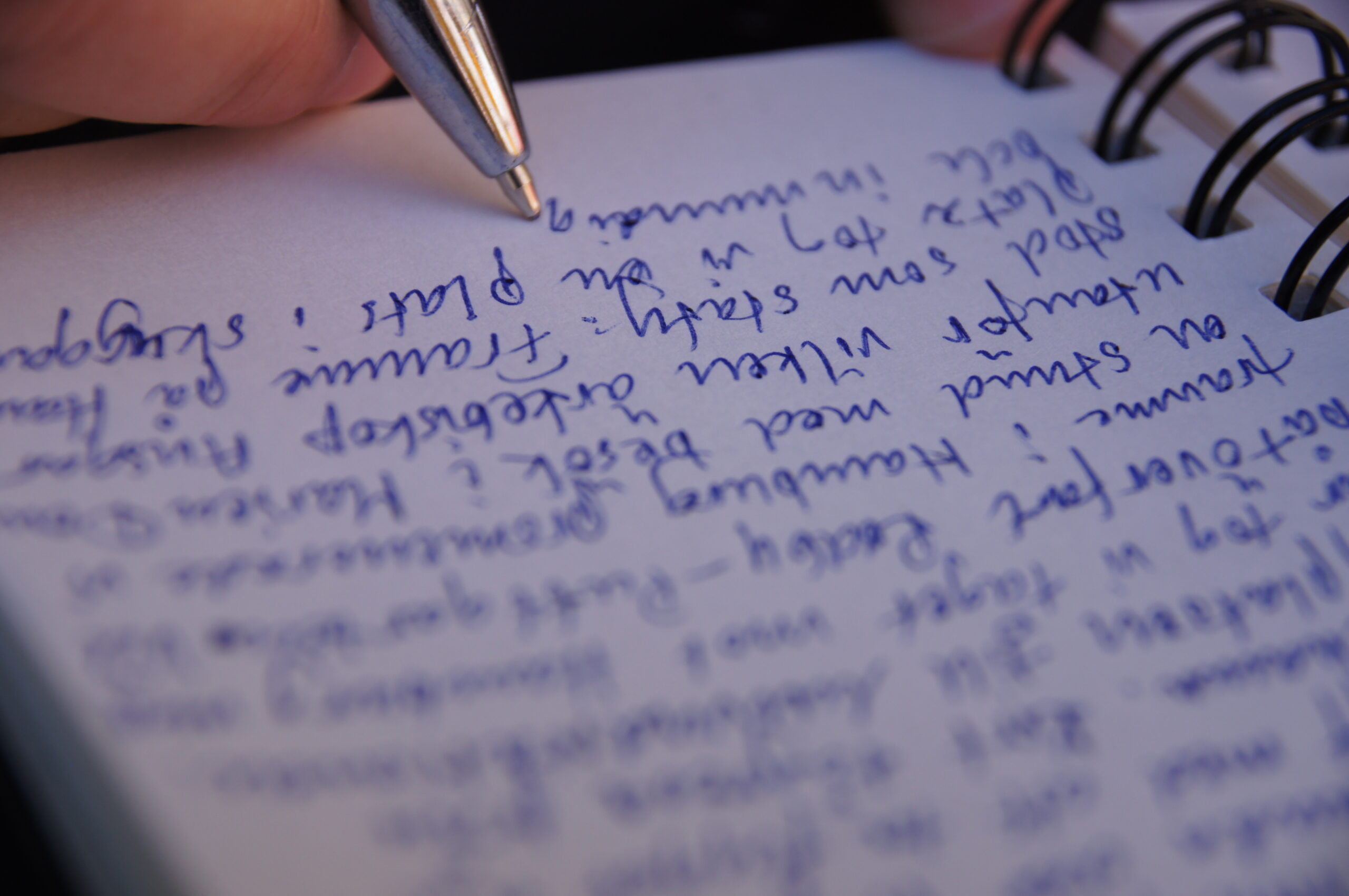 Foto com o zoom de um bloco de notas cheio de coisas escritas em um idioma não identificável e uma mão segurando uma caneta